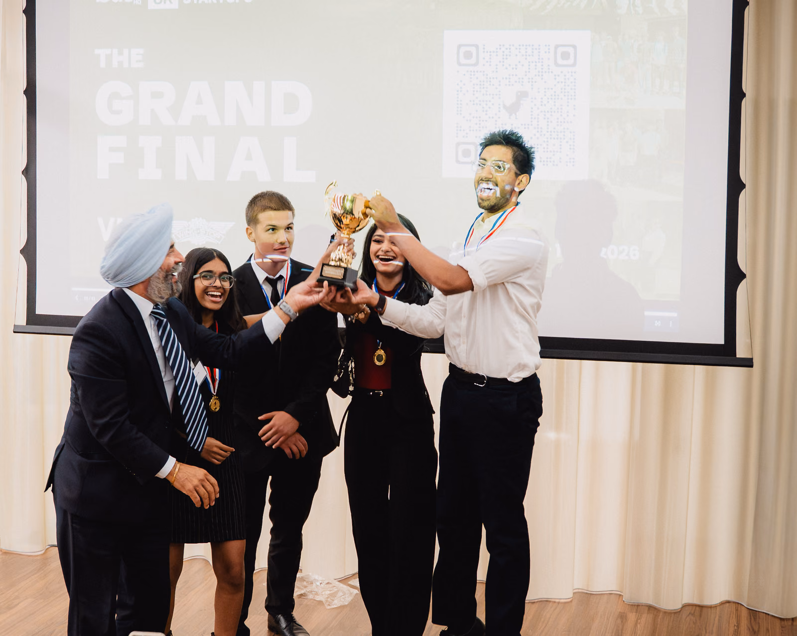 A team lifting a trophy in front of the Grand Final screen.